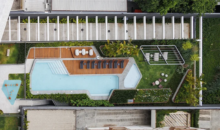 foto aérea de uma área de lazer. No centro, está uma piscina. De um lado, o deck de madeira onde ficam as espreguiçadeiras. Nos outros lados, os jardins do paisagismo><span height=
