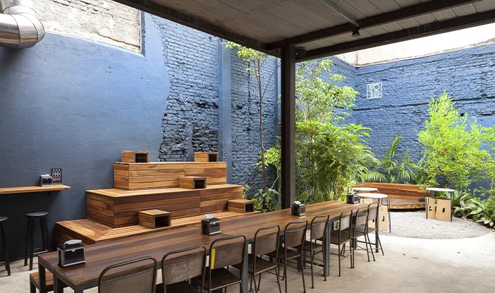 Pátio da Void, um estabelecimento em São Paulo. Na foto, há uma grande mesa de madeira ao centro, e, no fundo, uma parede azul. À direita, uma pequena área livre, com bancos em madeira também e vegetação no entorno<span height=