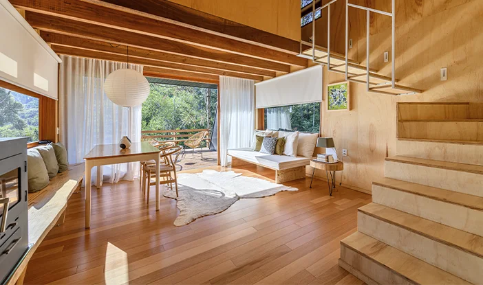 Área social de uma casa toda em madeira. Em um primeiro plano, estão as escadas. No segundo plano, as salas de jantar e estar