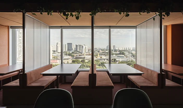 Foto de mesas com bancos estofados vermelhos, separadas por divisórias de vidro. No fundo, grandes janelas de vidro permitem um vista de toda a cidade><span height=