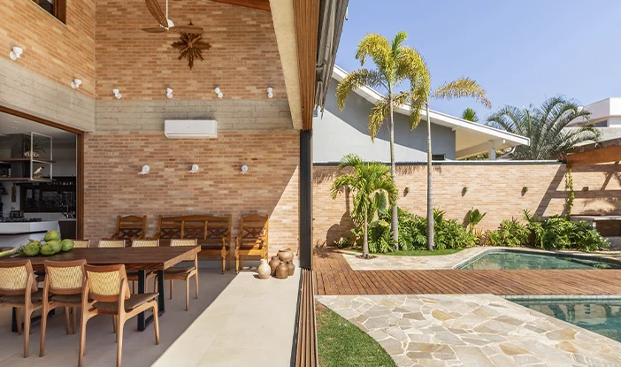 área de lazer da casa lima, com abertura direta para a piscina. à esquerda, está a varanda gourmet, com cadeiras e uma grande mesa de madeira. No centro, há trilhos para as portas de vidro de correr. à esquerda, está a passarela de madeira que leva à piscina e passa sobre ela<span height=