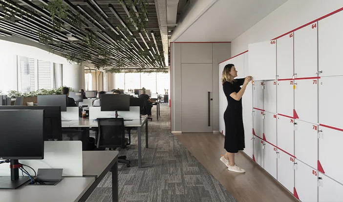 Foto do workspace da Metso com armários brnacos ocupando a parede à direita. à esquerda, estão as baias de trabalho com mesas brancas e cinzas. No teto, há várias luminárias com plantinhas pendentes.<span height=