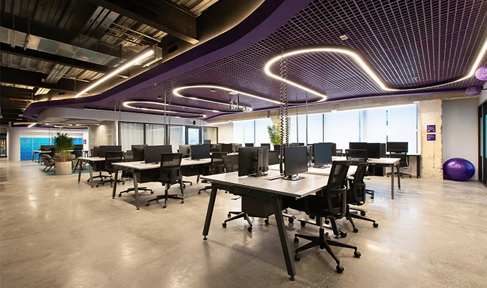 foto de um espaço de trabalho na nova sede do Nubank. Há baias de trabalho, cadeiras pretas, iluminação branca e grids metálicos na cor roxa no teto