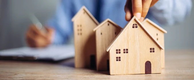 foto de uma casa de madeira em miniatura, e uma pessoa atrás que, com uma mão segura uma caneta, e com a outra segura a ponta da casinha  