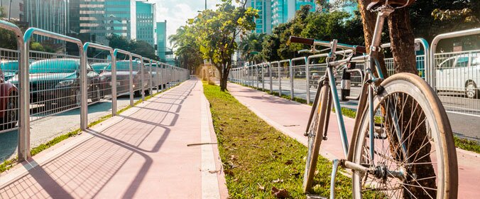 foto de uma bicicleta apoiada em uma árvore que está no canteiro central de uma ciclovia com duas mãos 