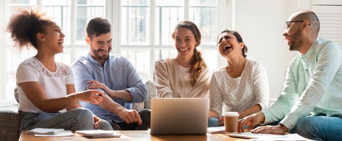 cinco pessoas reunidas tomando café em volta de um notebook e rindo