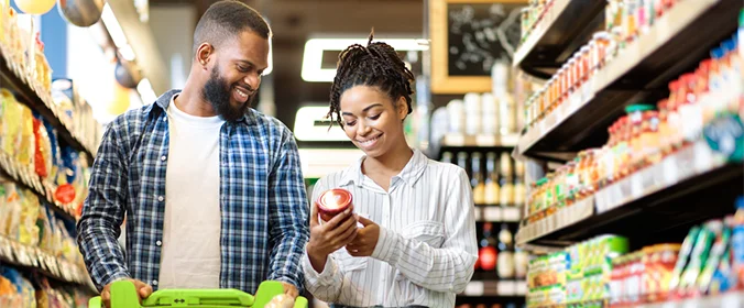 foto de duas pessoas em um mercado, com um carrinho a frente, e a mulher segurando um molho de tomate