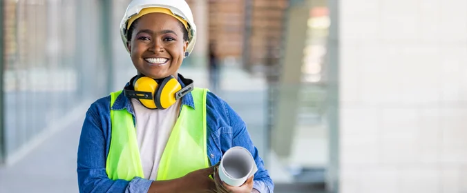 foto de uma mulher que sorri para a câmera, com traje de segurança de canteiros de obra, capacete e bloqueadores de ruídos para ouvidos  