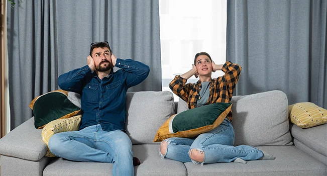 foto de duas pessoas em um sofá tampando os ouvidos com as mãos e olhando para cima