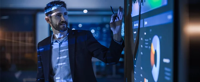 homem em uma sala escura com o projetor ligado aponto para uma tela grande com uma caneta na mão, como se estivesse fazendo uma apresentação