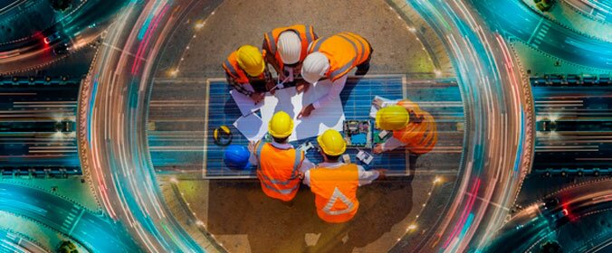 foto de uma vista superior, em que trabalhadores com roupas de proteção de canteiros de obras se reunem sobre uma mesa e ao fundo aparecem luzes sobre uma estrada que dão a sensação de movimento