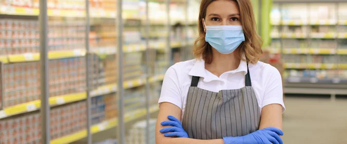 mulher com máscara, avental e luvas, de braços cruzados, em um supermercado