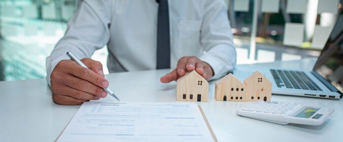 imagem de um homem em um escritorio segurando miniaturas de casas em uma mao e uma caneta em cima de um documento na outra