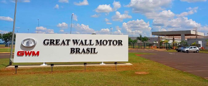 foto da placa da fábrica da GWM, com o nome e logo da empresa. Tem um estacionamento ao fundo, com dois carros estacionados. Tudo ao ar livre e com pouca vegetação atras da placa, a nao ser grama