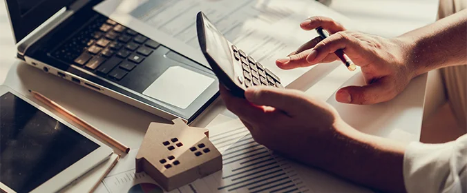 foto de mãos segurnado uma calculadoras com papéis e miniaturas de casas ao fundo