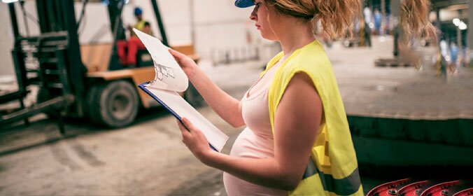 foto de uma moça gravida com equipamentos de seguranca de uma obra e analisando uma prancheta. No fundo, uma industria desfocada