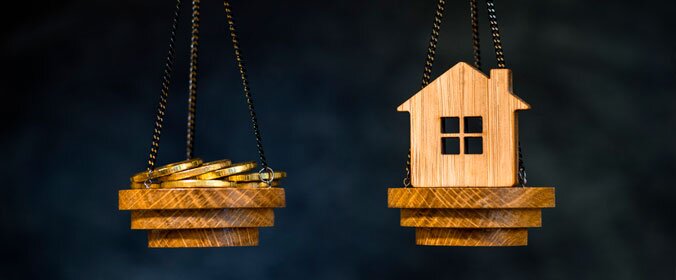 foto de uma balança pequena, de madeira. De um lado, uma mini casinha de brinquedo. Do outro, algumas moedas. Elas se equilibram