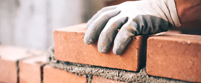 foto de uma mão, com luvas, segurando um tijolo e colocando-o sobre uma parede com concreto