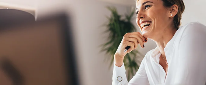foto de uma mulher com uma caneta na mão, sentada na ponta de uma mesa e sorrindo