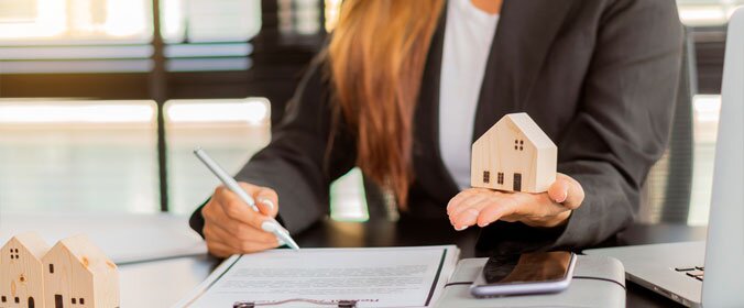 mulher atrás de uma mesa assinando um contrato com uma mãe e segurando uma miniatura de casa em outra mão