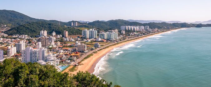 imagem aerea de itajai, em santa catarina, que evidencia a praia, o mar, a cidade com varios predios e montanhas ao fundo