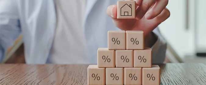 foto de uma pessoa colocando um quadradinho com o desenho de uma casa sobre uma pirâmide de outros quadradinhos, que, por sua vez, possuem o desenho de porcentagem