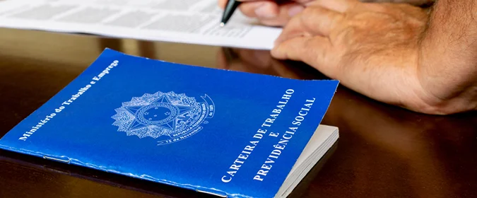 foto de uma carteira de trabalho e uma pessoa com uma caneta na mao ao fundo