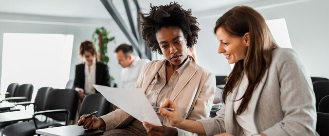 imagem de duas mulheres conversando e lendo um papel. O fundo está desfocado, mas existem outras duas pessoas em bancos atrás delas que também estão conversando sozinhos