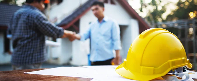 foto de um capacete de proteção, utilizado em canteiros de obra. Ao fundo, desfocados, dois homens se cumprimentam com um aperto de mão.