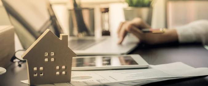 foto da  mão de uma pessoa mexendo em um notebook e uma casa em miniatura em frente