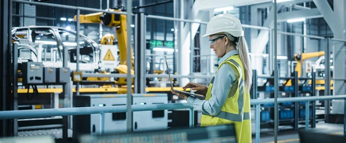 foto de uma mulher, vestida com equipamentos de segurança e mexendo em um notebook, dentro de uma indústria