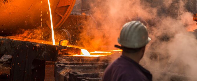 imagem de um trabalhador industrial de costas, protegido com os devidos equipamentos, e uma fornalha liberando fumaça ao fundo