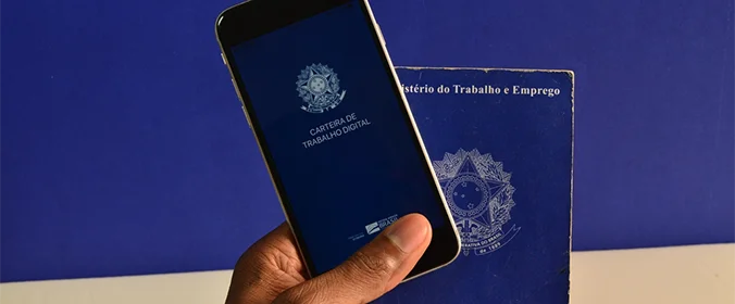 foto de uma mão segurnado um celular, que exibe o logo da carteira digital, e uma carteira digital física, com um fundo azul