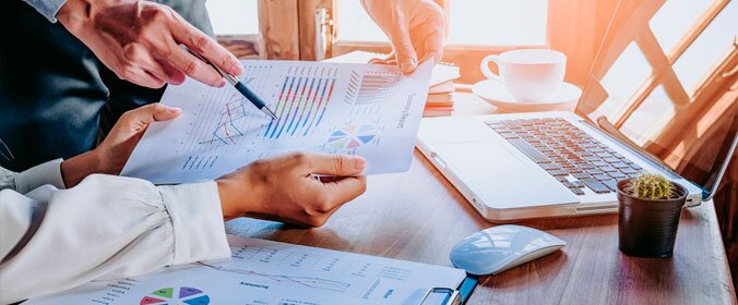 imagem de uma reunião em que aparecem duas pessoas, sendo que uma delas segura um papel com gráficos e dados e a outra se inclina sobre o papel com uma caneta, como se estivesse explicando. Ao fundo, um notebook, uma xícara de café e um vaso pequeno de planta