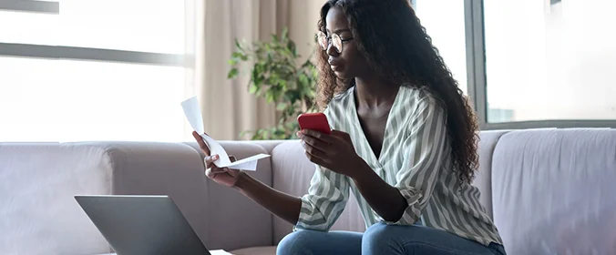 foto de uma mulher sentada em um sofá, olhando para um notebook, segurando um papel em uma mão e um celular na outra mão