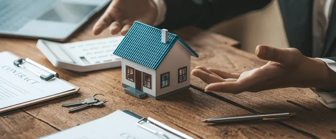 pessoa sentada e na messa estão: miniatura de uma casa, chaves, calculadora, canet a e papel com o título "contrato"