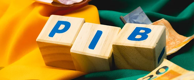 foto de três cubos de madeira, com letras que formam a palavra PIB. Eles estão sobre uma bandeira verde e amarela, com algumas cédulas de real em volta