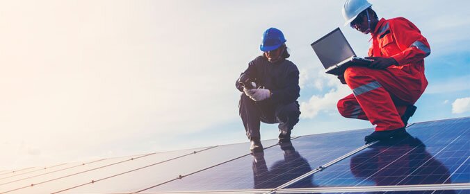 dois homens, com uniforme característico de canteiros de obras, estão sobre várias placas solares e segurando um notebook