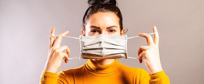 mulher de cabelos presos e blusa amarela prestes a colocar a mascara no rosto, presa as orelhas