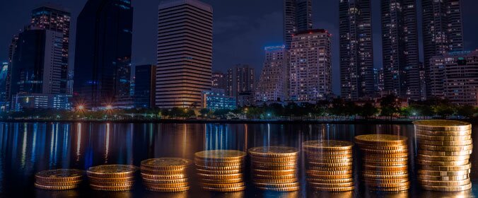 foto de uma cidade a noite iluminada ao fundo, e, na frente, varias pilhas de moedas. Cada nova pilha à direta tem uma moeda a mais.