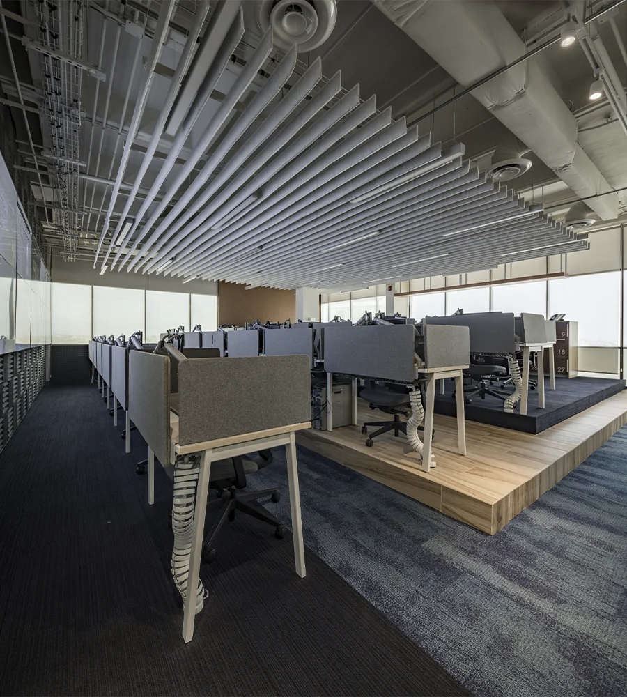 sala comercial do escritório Execon com carpete em azul marinho e dois tablados de madeira. Sobre os tablados, uma série de computadores nas mesas são separadas por baias na cor cinza. Em cima, a luminária é constituída por várias tiras brancas